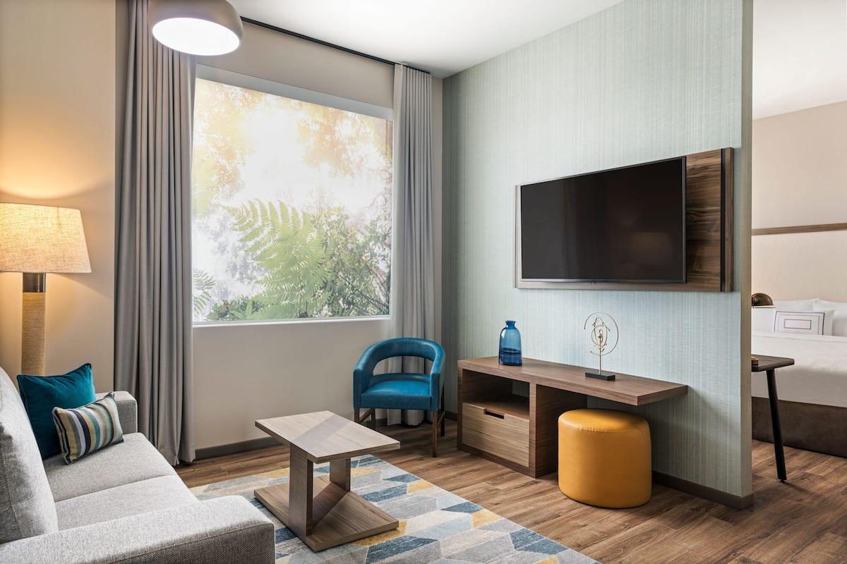 A modern hotel room with a sofa, a desk, a TV, and a large window with curtains.