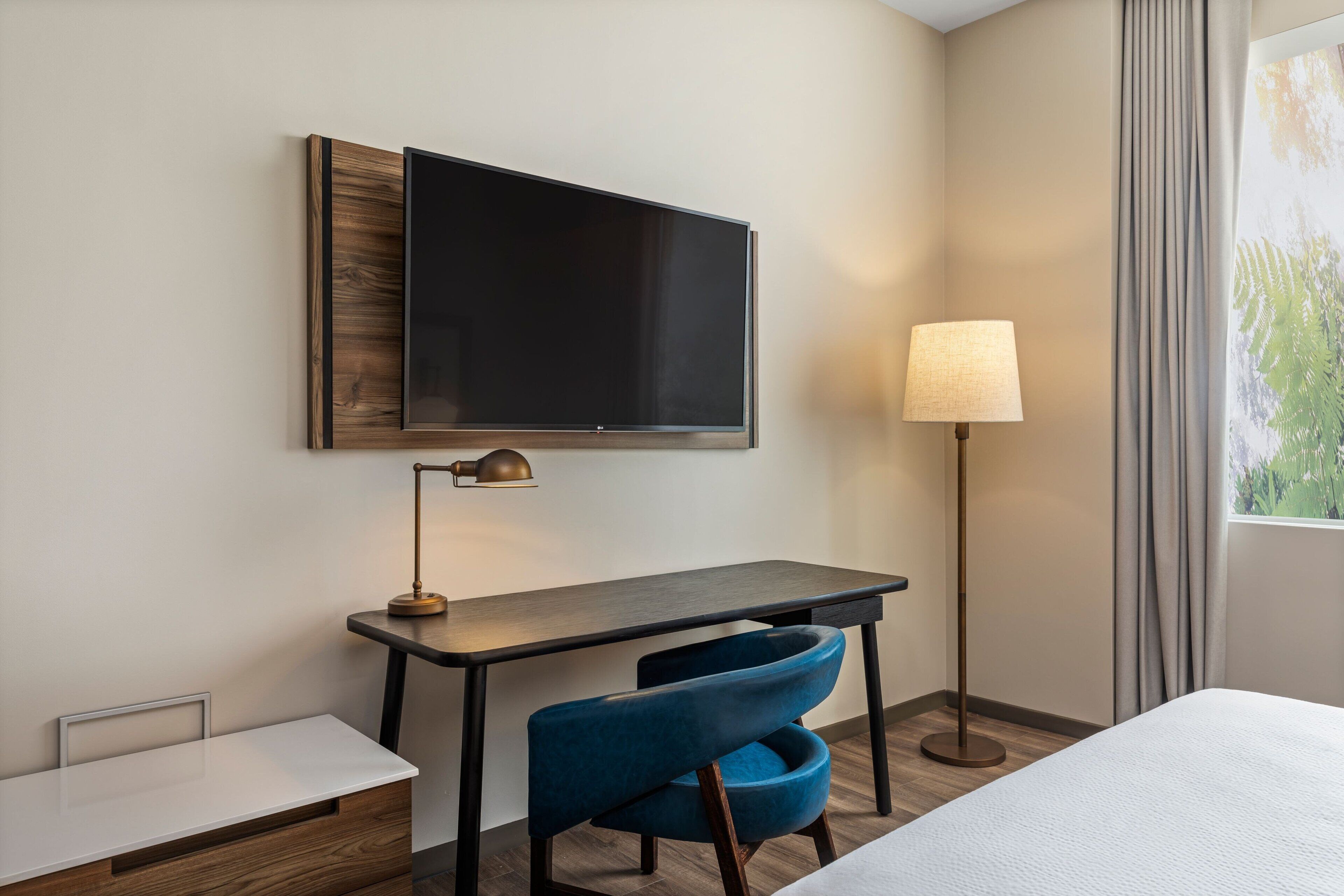 Une chambre d’hôtel équipée d’un téléviseur à écran plat fixé au mur, d’un bureau avec une lampe, d’une chaise bleue et d’un lit.