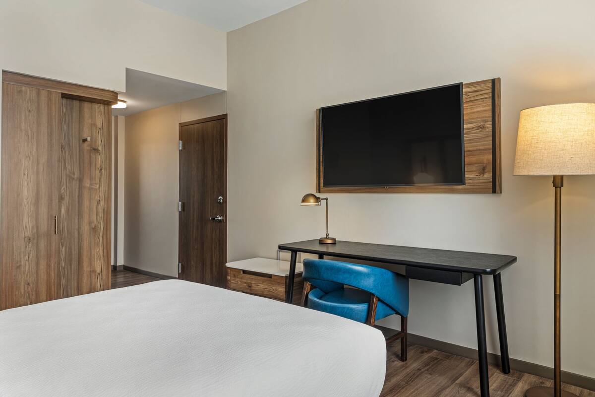A hotel room with a bed, a desk with a chair, a TV mounted on the wall, and a floor lamp.