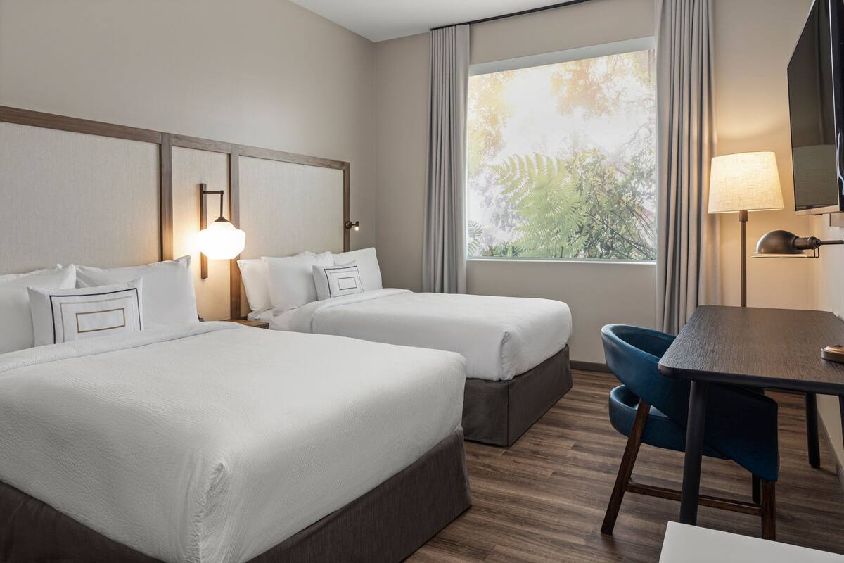 A hotel room with two beds, a desk, a chair, and a large window with curtains.