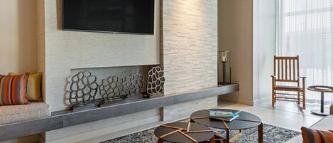 A modern living room with a flat-screen TV mounted on a textured wall, a built-in fireplace with decorative items, a wooden chair, and a round coffee table.