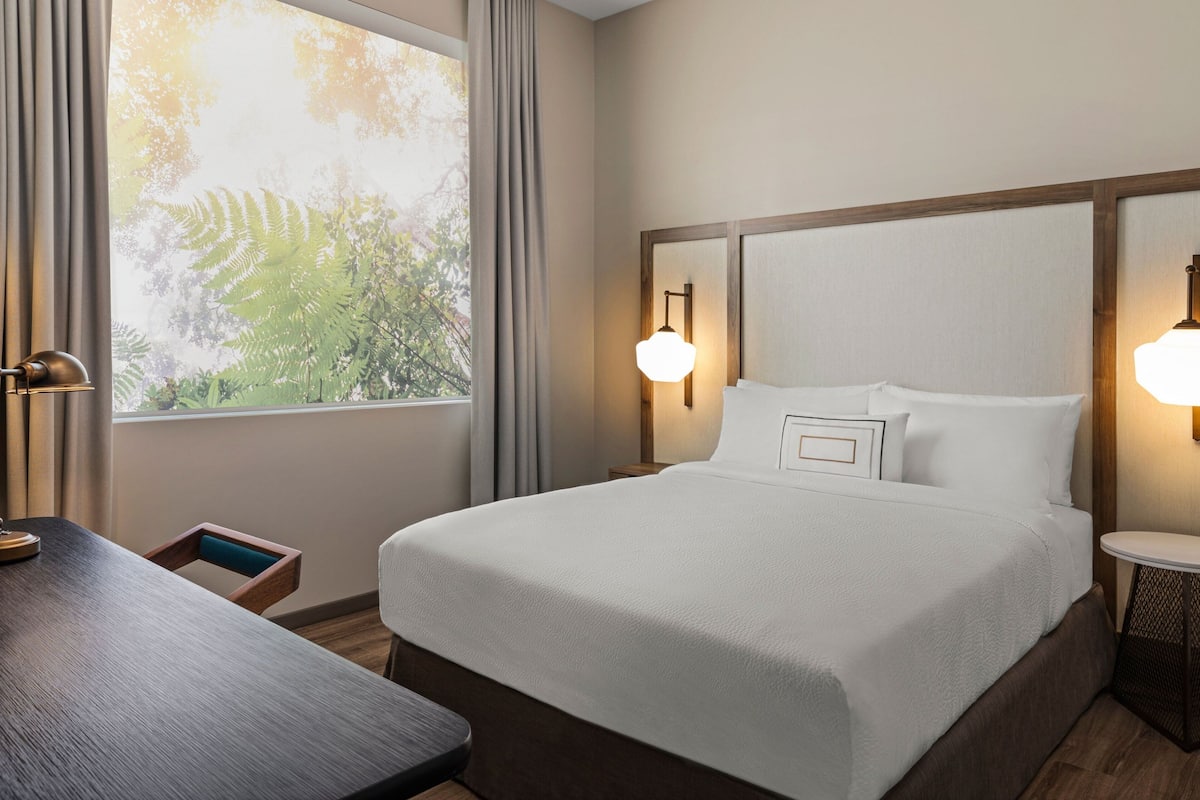 A hotel room with a large bed, two bedside lamps, a small table, and a window with curtains.