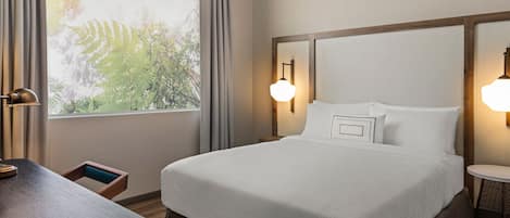 A hotel room with a large bed, two bedside lamps, a small table, and a window with curtains.