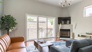 A modern living room with a leather sofa, grey armchair, and a coffee table. There is a fireplace, a TV mounted on the wall, and a large window with a view of the outside. - Greystone, 3500 sq ft, Rooftop Access, Sleeps 16 (Chicago)