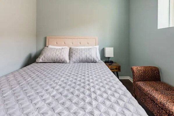 Una cama bien tendida con un cabecero acolchado, una mesita de noche con una lámpara y un sillón estampado de leopardo.