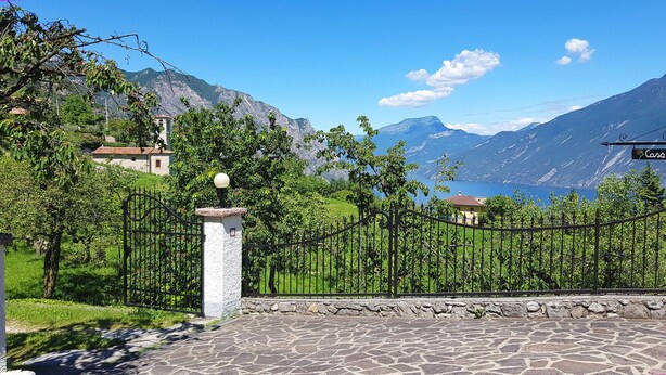 A stone-paved entrance with a metal gate, a white pillar with a spherical light, and a view of a lake with mountains in the background.