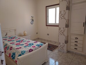 Un dormitorio con cama, mesita de noche, lámpara, reloj de pared, ventana y armario.