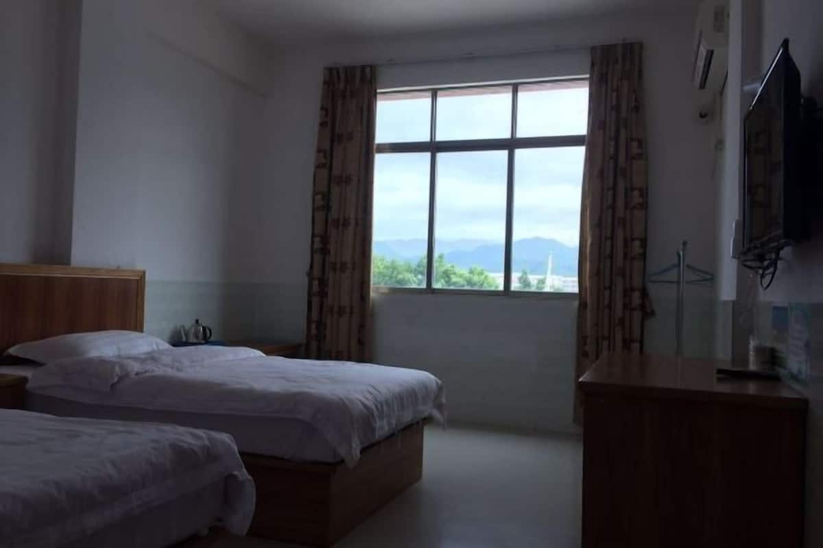 A hotel room with two beds, a wooden headboard, a desk, a television, and a window with a view of mountains.