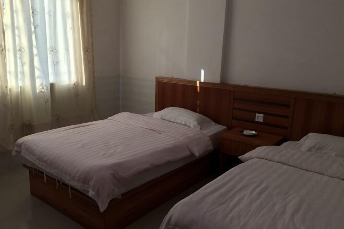 A hotel room with two beds, wooden headboards, a bedside table with a lamp, and a window with sheer curtains.