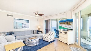 A living room with a sofa, ottoman, TV, and a view of the ocean through large windows.