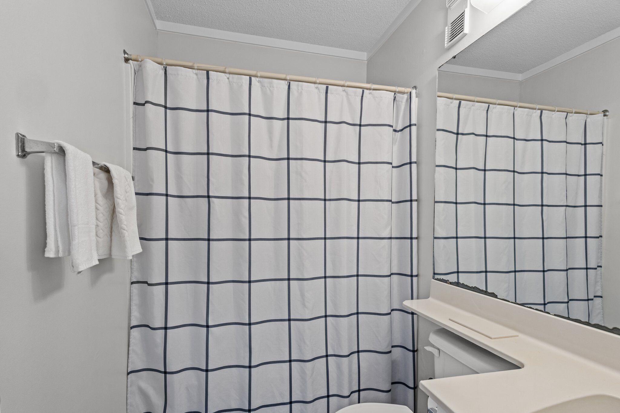 A bathroom with a white and blue checkered shower curtain, a white towel hanging on a metal towel rack, and a white sink with a mirror above it.
