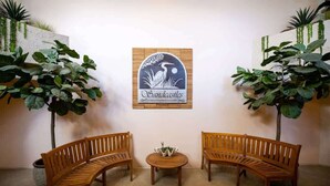 A room with two wooden benches facing each other, a small round table with a plant, and a wall-mounted sign with a bird and the word "Sandcastles". - Sandcastle Court 219 (Nassau County)