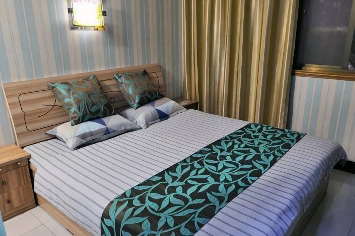 Een net opgemaakt bed met gestreept beddengoed en een plaid met bladmotief, een houten nachtkastje en een wandlamp.