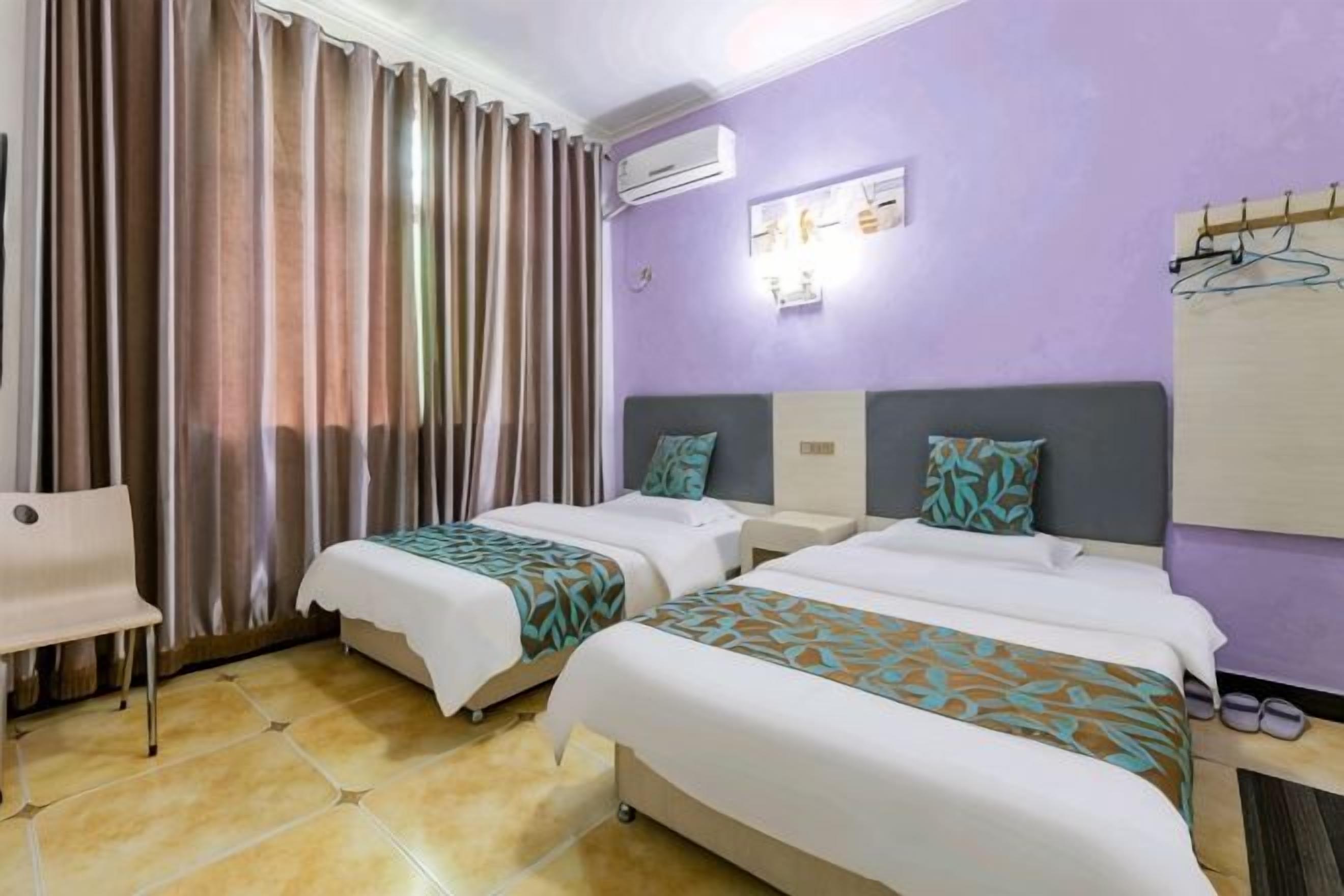 A hotel room with two beds, a chair, an air conditioner, and a wall-mounted light fixture.