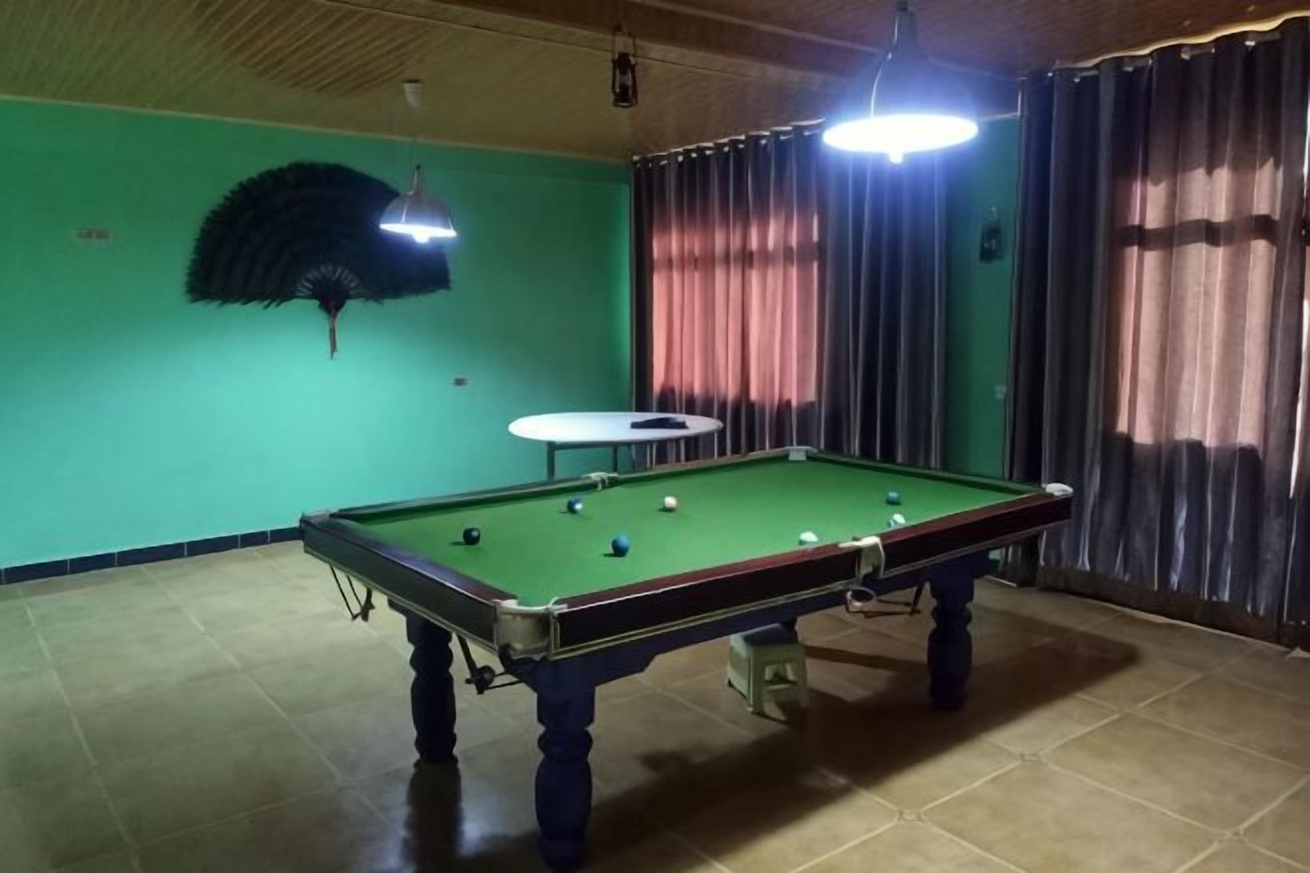 Una mesa de billar en una habitación con paredes verdes, un ventilador de techo y cortinas.