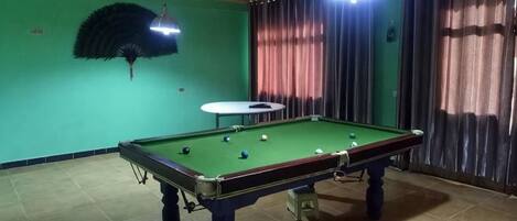 Una mesa de billar en una habitación con paredes verdes, un ventilador de techo y cortinas.