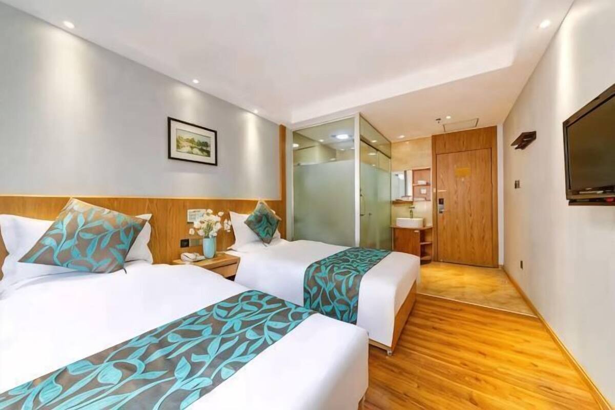 Een hotelkamer met twee bedden, een houten vloer, een televisie en een zichtbare badkamer door een glazen deur.
