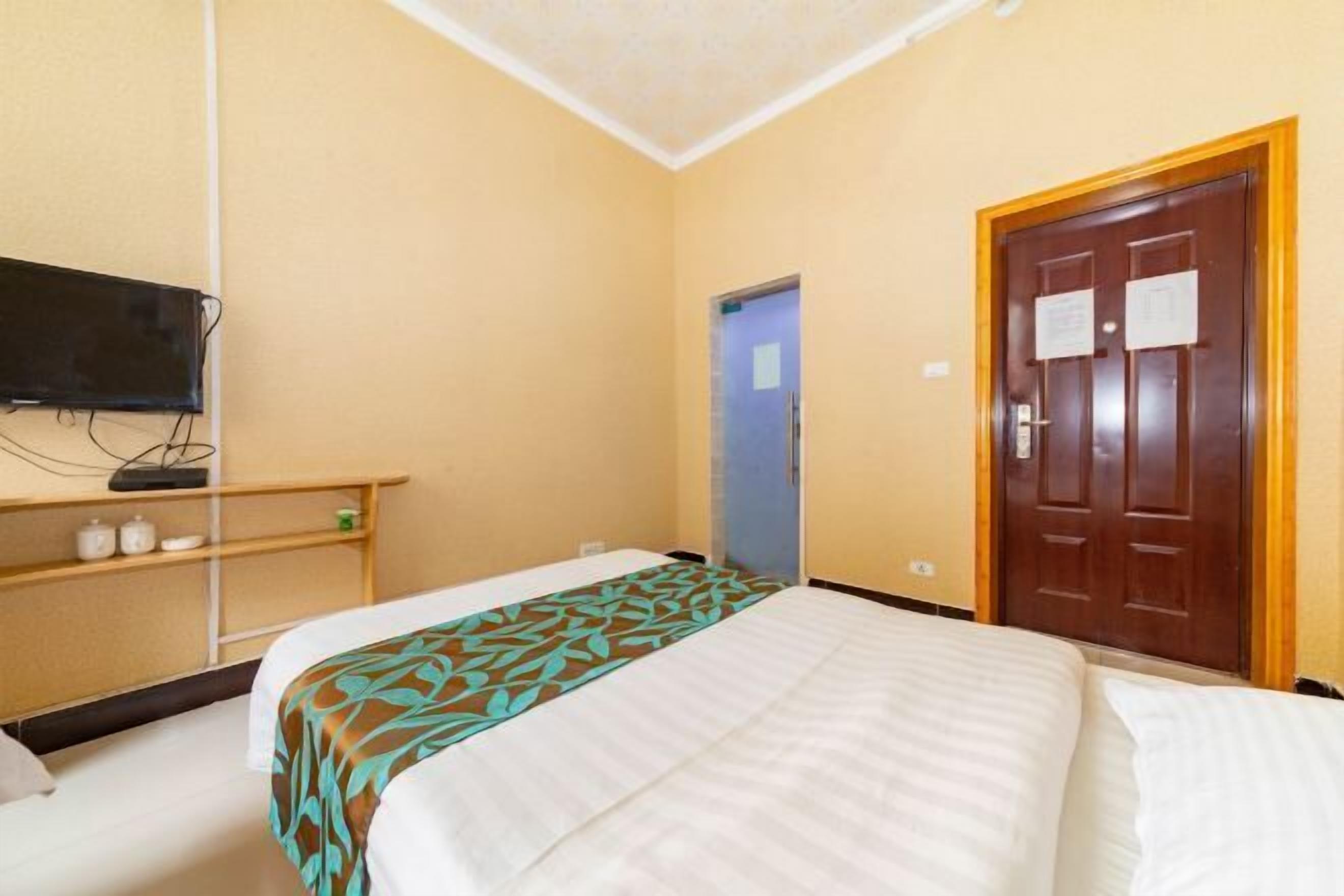A hotel room with a bed, a television on a stand, a door, and a shelf with items.