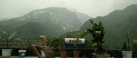 A rooftop terrace with potted plants, a wicker chair, and a mountain backdrop.