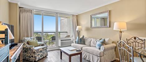 A living room with a sofa, a chair, a coffee table, a TV, and a balcony with a view.