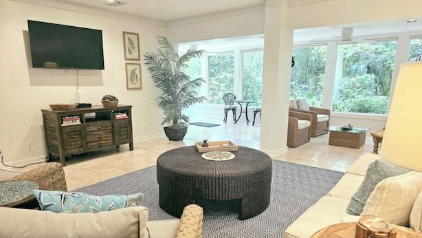 A spacious living room with a round ottoman, a flat-screen TV, wicker furniture, and a large window offering a view of greenery. - Fairway Oaks 3209 (Fernandina Beach)