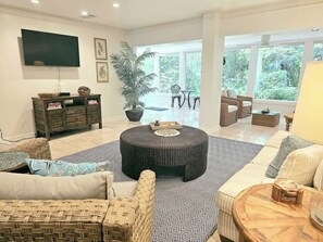 A spacious living room with a round ottoman, a flat-screen TV, wicker furniture, and a large window offering a view of greenery. - Fairway Oaks 3209 (Fernandina Beach)