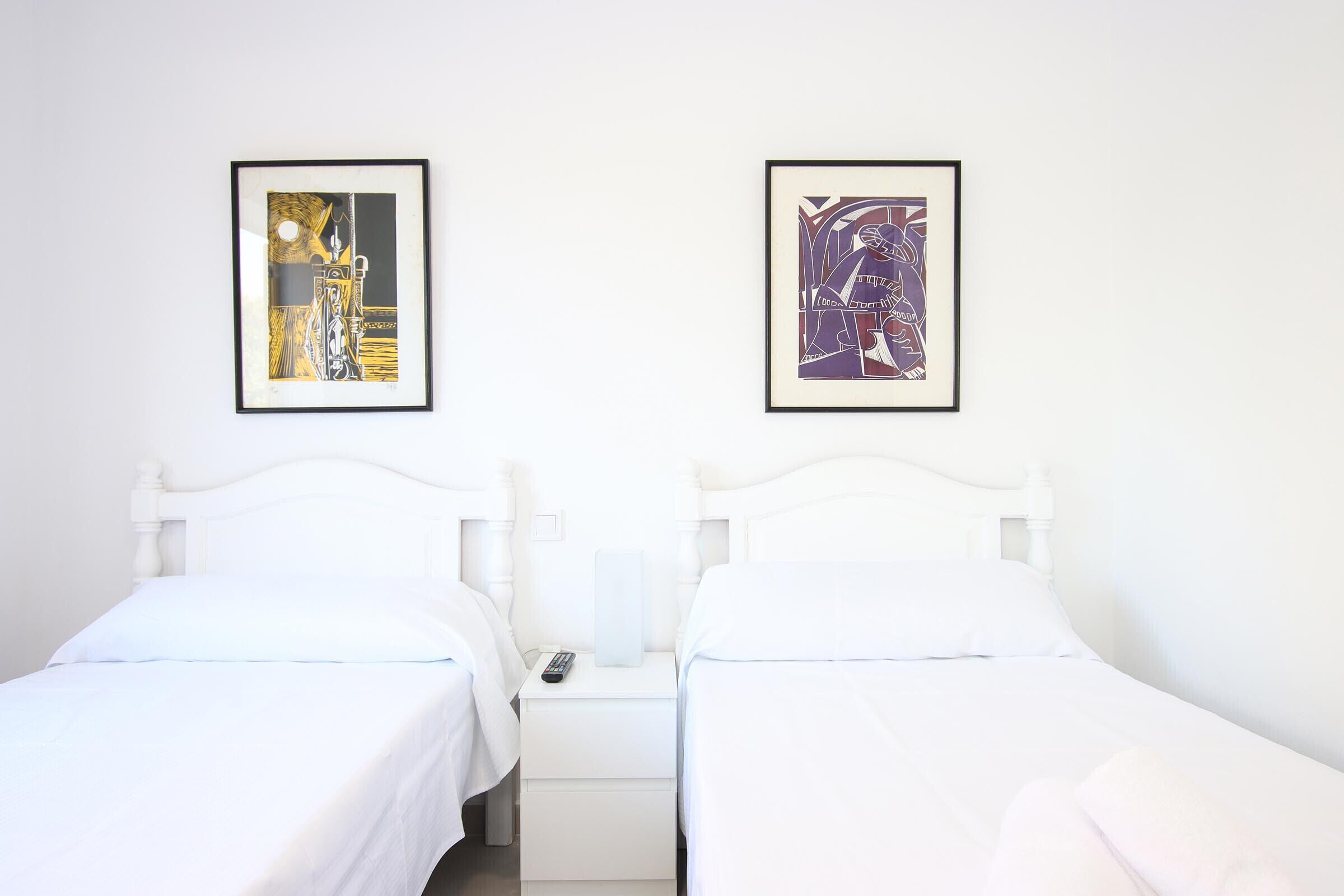 Dos camas individuales con ropa blanca, una mesita de noche con control remoto y dos cuadros en la pared.