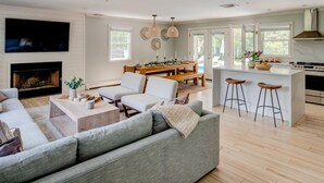 A modern living room with a large sofa, a coffee table, and a fireplace. There is a dining area with a wooden table and chairs, and a kitchen with a stove and bar stools. - Re-modernized home with heated saltwater pool, home office, gym, electric... (Southampton)