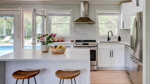 A modern kitchen with white cabinetry, a central island with stools, a gas stove, and a stainless steel refrigerator. - Re-modernized home with heated saltwater pool, home office, gym, electric... (Southampton)