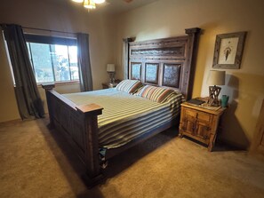 Une chambre à coucher avec un lit en bois, une table de chevet, une lampe, une fenêtre avec des rideaux et un tableau encadré au mur.
