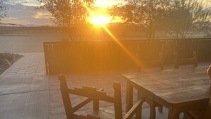 En træterrasse med et bord og stole, et metalhegn og en solnedgang i baggrunden.
