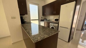 A kitchen with granite countertops, dark wooden cabinets, a stainless steel refrigerator, a washing machine, and a microwave.