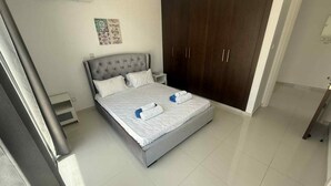 A neatly made bed with white linens and rolled towels, a dark wooden wardrobe, and a small bedside table with a lamp.