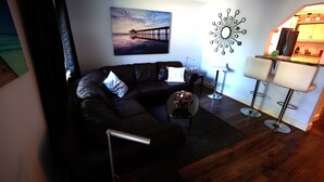 A living room with a dark leather sofa, a coffee table, a painting of a pier, a modern mirror, and a kitchen area with bar stools.