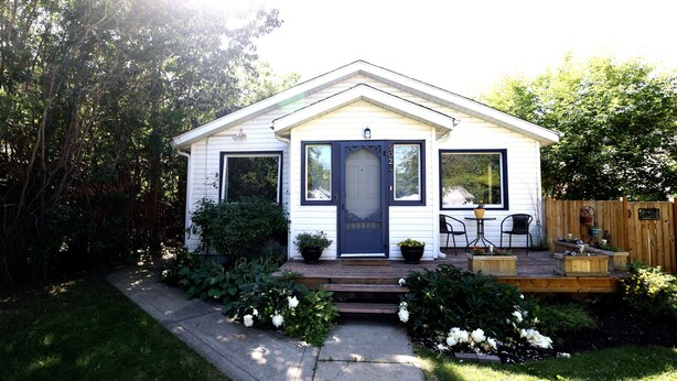A small, single-story house with a dark blue front door, white siding, and a small porch with two chairs and a table.