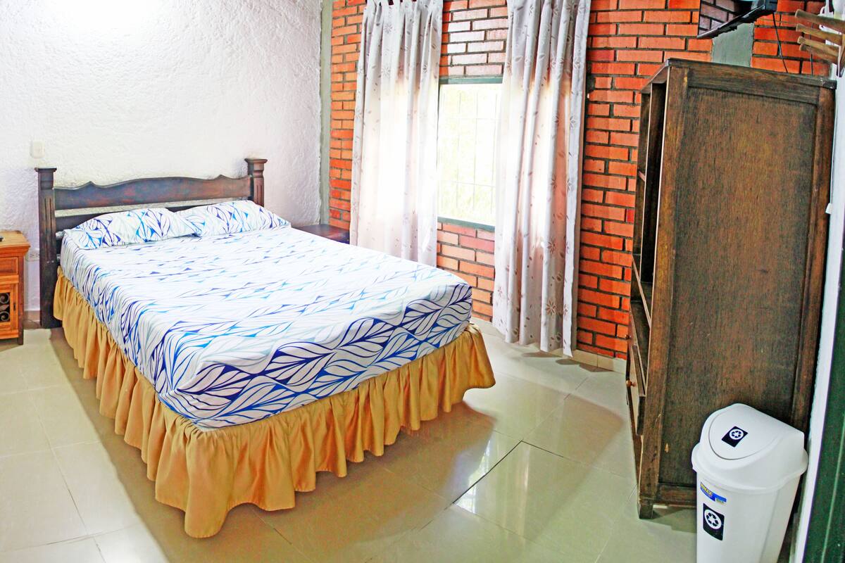 A bedroom with a bed, a wooden nightstand, a dresser, a mirror, a window with curtains, and a trash bin.