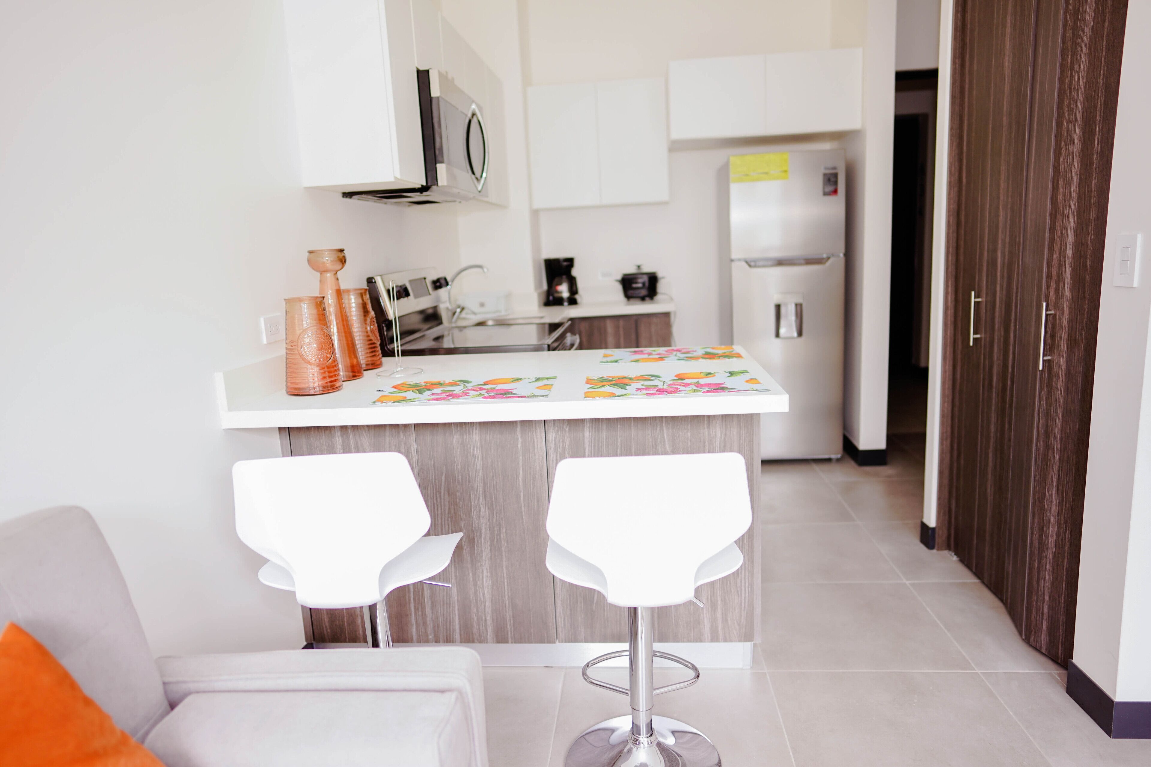 A modern kitchen with white cabinets, a stainless steel refrigerator, and a small island with two white bar stools.