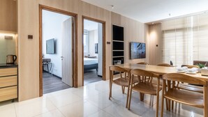 Et moderne hotelværelse med trægulv, et spiseområde med et træbord og stole, et fjernsyn monteret på væggen og et stort vindue med persienner.