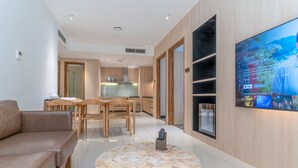 En moderne stue med sofa, et spiseområde med træstole, et køkken i baggrunden og et tv på væggen, der viser et program.