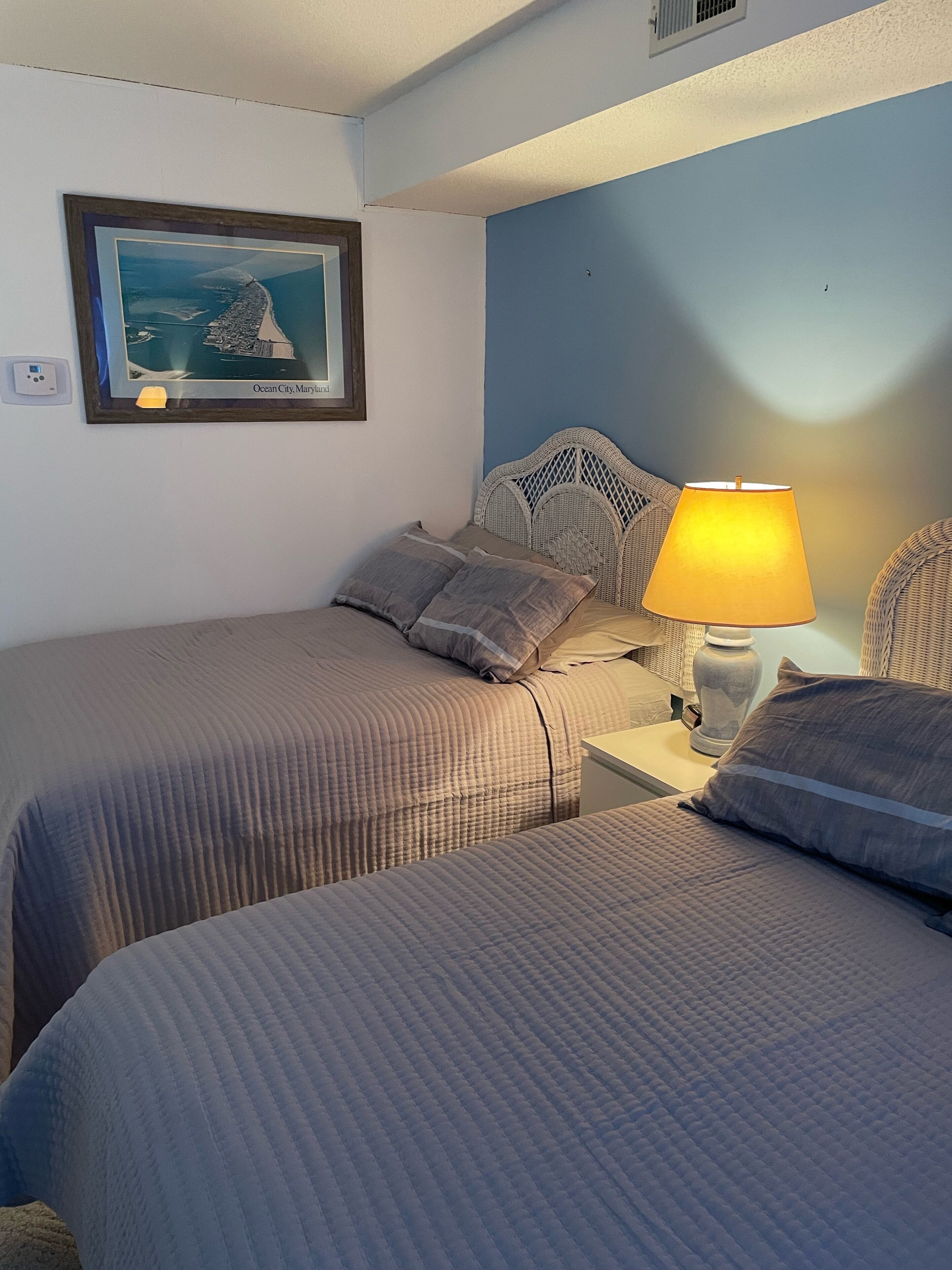 Une chambre d’hôtel avec deux lits, une table de chevet avec une lampe, un tableau encadré au mur et une prise de courant.