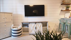Ein Wohnzimmer mit einem an der Wand befestigten Flachbildfernseher, einem Korbuscheinheit, einem gestreiften Ottomanen und einem Blumentopf.