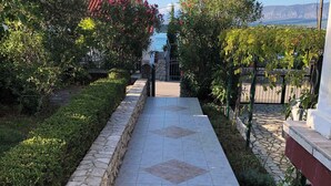 A paved pathway leading to a gated entrance, flanked by stone walls and lush greenery, with a clear view of a building and distant mountains.