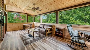 L'intérieur d'un chalet en bois comprend un canapé, une table basse, un fauteuil à bascule et une grande fenêtre donnant sur une forêt.