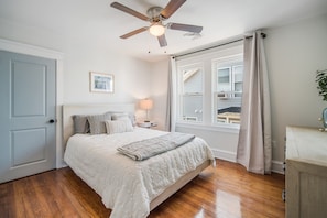 Quarto com cama, ventoinha no teto, mesa de cabeceira, abajur e janela com cortinas.