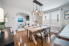 Uma sala de jantar moderna com uma mesa e cadeiras de madeira, um candelabro e vista para a sala de estar ao fundo.