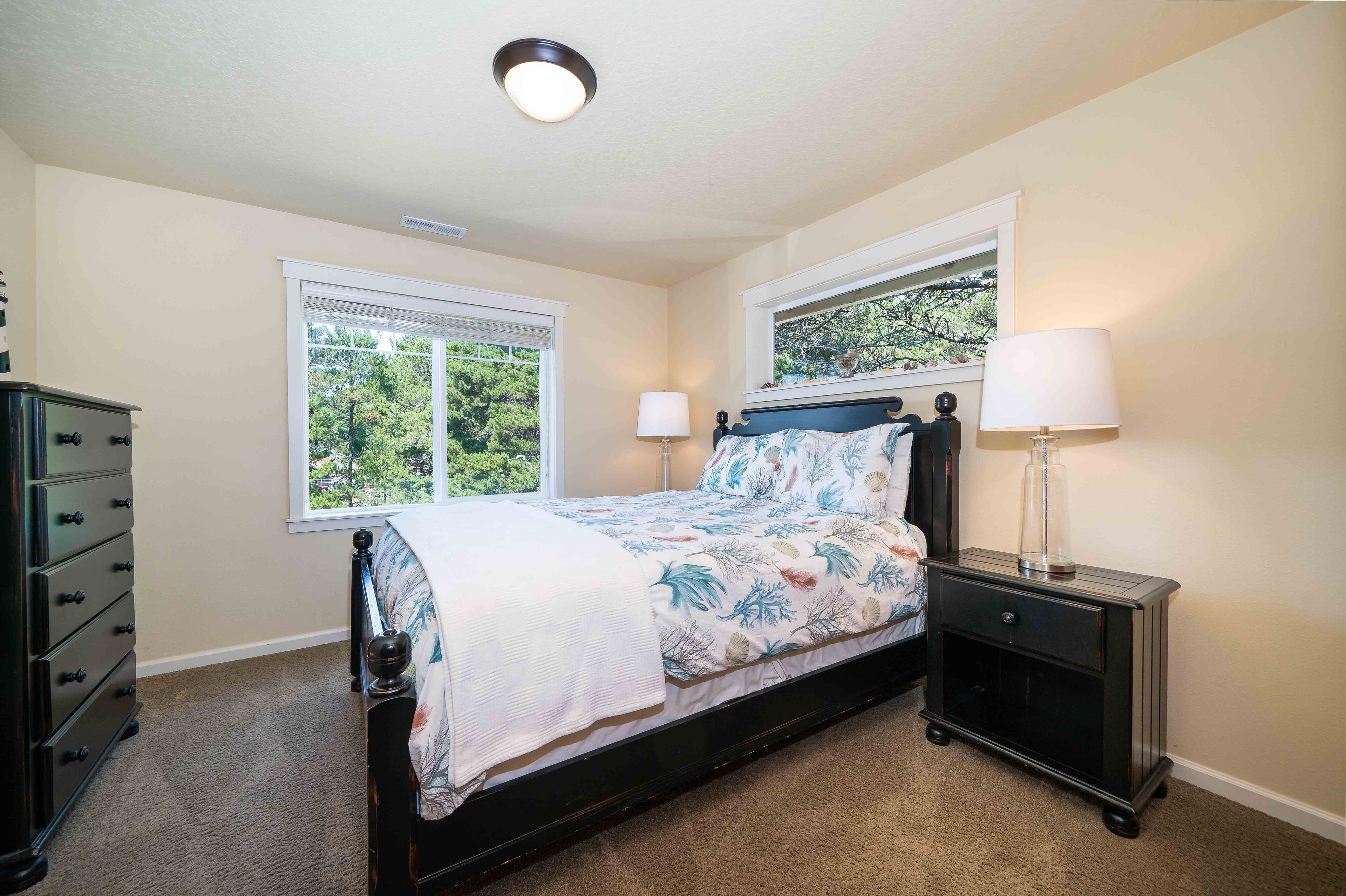 A bedroom with a bed, two windows, a dresser, and a nightstand with a lamp.