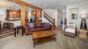 A spacious living room with a brown leather sofa, wooden coffee table, and wicker armchair. A kitchen with wooden cabinets and a refrigerator is visible in the background. - Spacious 2-Story Home, walk to the beach, fenced yard, patio, bbq and fire pit (Pacific City)