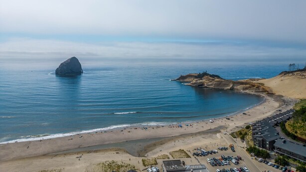 An aerial view of a beach with a large rock formation, sandy beach, and a parking area with cars. - Spacious 2-Story Home, walk to the beach, fenced yard, patio, bbq and fire pit (Pacific City)