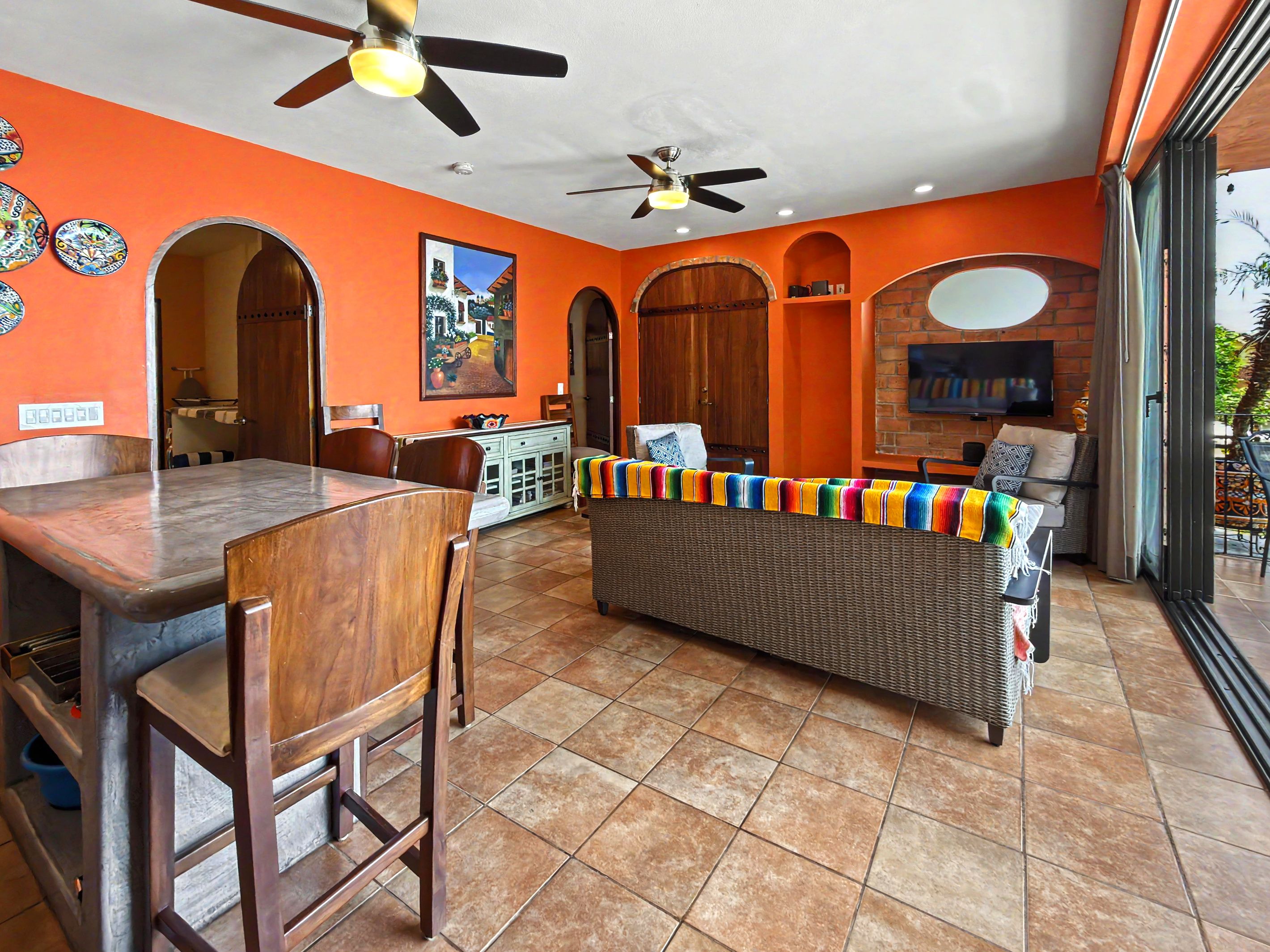 A dining area with a wooden table and chairs, a sofa with a colorful blanket, and a TV mounted on a brick wall.