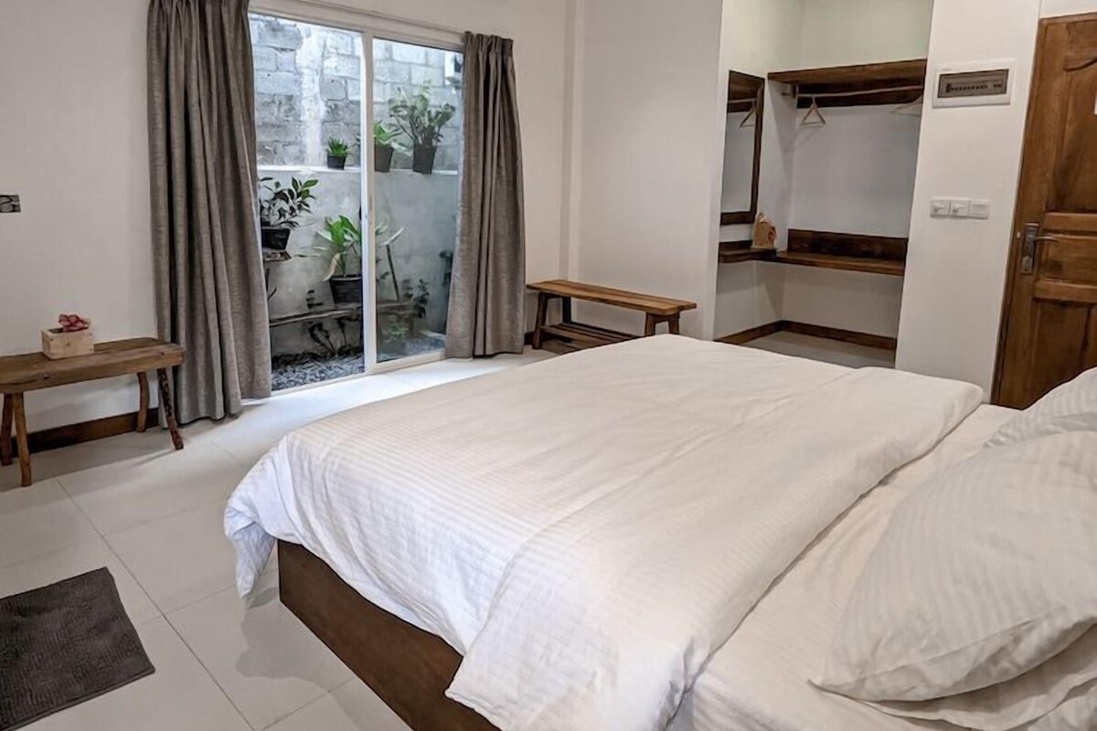 Un dormitorio con una cama grande, ventilador de techo, equipo de aire acondicionado, una banca y un ventanal corredizo que da a un área exterior con plantas.